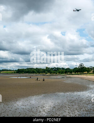 Dog walkers on wet sand at low tide with BA plane overhead, Cramond, Edinburgh, Scotland, UK with British Airways plane flying in sky overhead Stock Photo