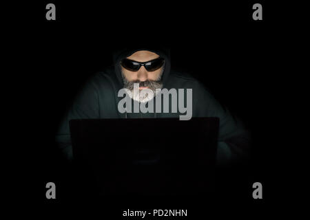 Portrait of a bearded computer hacker in a hoodie using laptop in a dark room. Stock Photo
