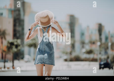 Rear view of young woman walking on city street wearing hat enjoying happy pleasant moment of her vacations Stock Photo