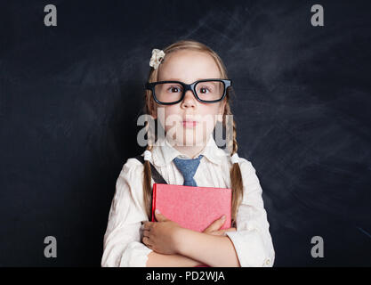 Smart Child on Blackboard Background. Happy little girl in glasses with red book. Back to school Stock Photo