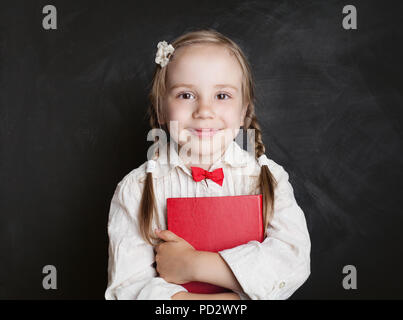 Cute child girl with book having fun on chalkboard background in elementary school classroom. Back to school, reading and education concept Stock Photo