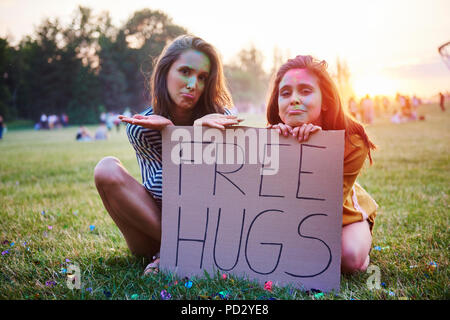 Young women sitting on grass holding free hugs sign at Holi Festival, portrait Stock Photo