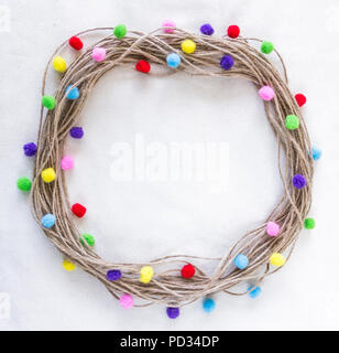 Hessian, twine, jute string wreath with multicolor, fluffy pom poms on raw cotton fabric ecru background Stock Photo