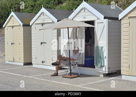 Man sitting in a chair reading outside a beach hut. Bournemouth, Dorset, UK, Monday 6th August 2018. Hot weather on the south coast beach as the glorious summer heatwave continues. Stock Photo