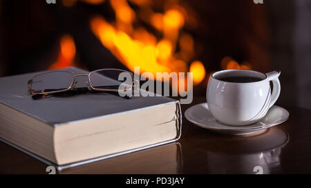 White cup of tea or coffee, glasses and old book near fireplace on wooden table. Winter and Christmas holiday concept Stock Photo