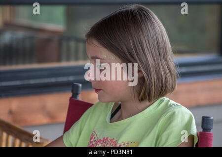 Pretty 8 year od girl, outside,  wearing makeup, gold glitter, sitting looking down, camera left, in somber mood. Stock Photo