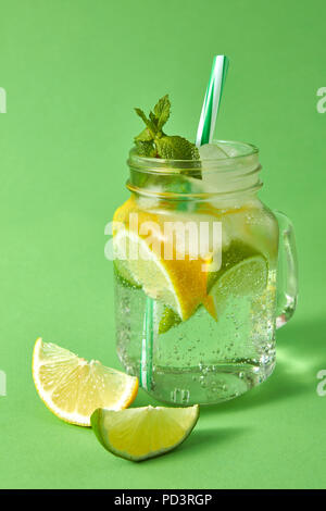 Homemade sparkling lemonade with ice, slices of lime and lemon, leaf of mint with plastic straws in a mason jar on a green background. Top view. Stock Photo