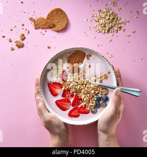 Healthy smoothie in white bowl with natural fruits, oat flakes and biscuits with woman's hands holding a bowl on pink background. Superfoods, natural  Stock Photo