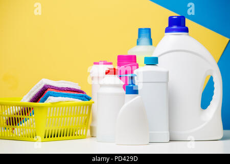 Photo of several bottles of cleaning products and multi-colored towels in basket isolated on blue, yellow background Stock Photo