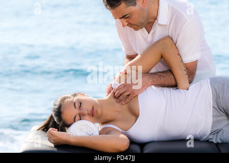 Close up portrait of male osteopath doing shoulder therapy on woman outdoors next to sea side. Stock Photo