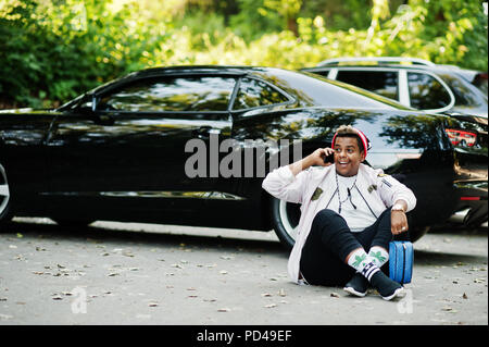 Stylish hipster arab man guy in hat posed outdoor in street against his black muscle car and speaking on mobile phone. Stock Photo