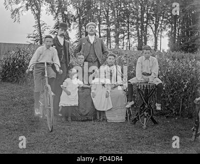 Alate Victorian photograph of a family posing for the camera, somewhere in England. There are two men and their wives, two young children, and two boys. One of the boys is sitting on a bicycle of the time. Stock Photo