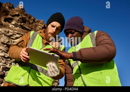 Low angle portrait of two industrial workers wearing reflective jackets, one of them African, inspecting mineral mines on worksite outdoors and using  Stock Photo