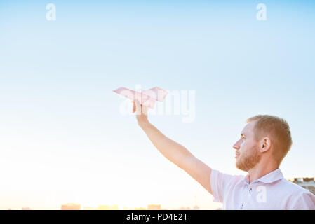 Man's hand holding paper airplane in blue sky at sunset light with city landscape background. Dreams and freedom concept. Selective focus. Copyspace Stock Photo