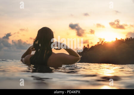 young woman from behind in indian ocean bathing and holding her hair during orange sunset with romantic mood Stock Photo