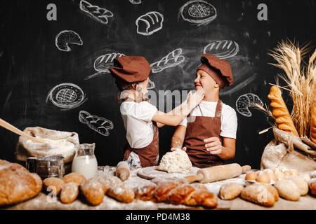 Adorable girl with brother in aprons on table with bread loaves making fresh dough and having fun Stock Photo