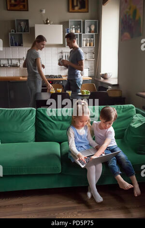 Kids using laptop together while parents in the kitchen Stock Photo