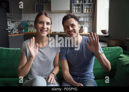 Smiling couple waving hands sitting on sofa looking at camera Stock Photo