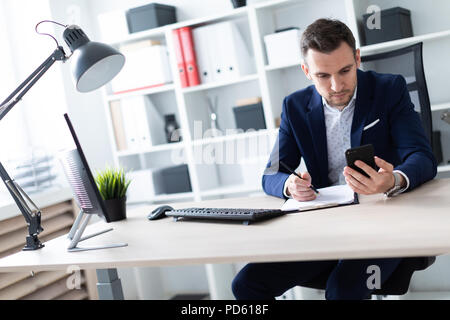A young man sits in the office at the table, looks into the phone and holds a pencil in his hand. The young man has documents. Stock Photo