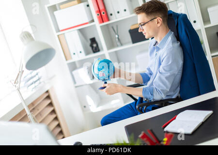 A young man sits in the office at a computer desk and holds a globe in his hands. Stock Photo