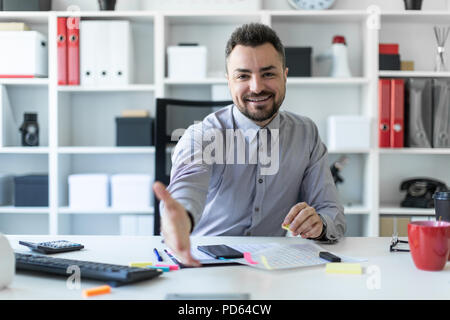 A young man sits at a table in the office and holds out his hand. The man is greeting someone. Stock Photo