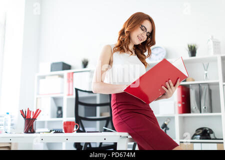 A beautiful young girl sat down on the table in the office and was holding a red folder. Stock Photo