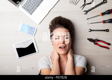 Stressed woman surrounded by assorted hand tools with her laptop, tablet and mobile phone as she lies on the floor in a close up on her face in a conc Stock Photo