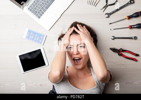 Stressed woman yelling with her hands to her hair as she lies on the floor surrounded by hand tool for repair of her digital devices in a close up on  Stock Photo
