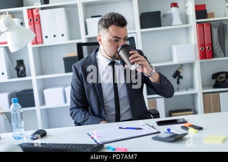 A man is sitting in the office at the table and drinking coffee from a glass. Stock Photo