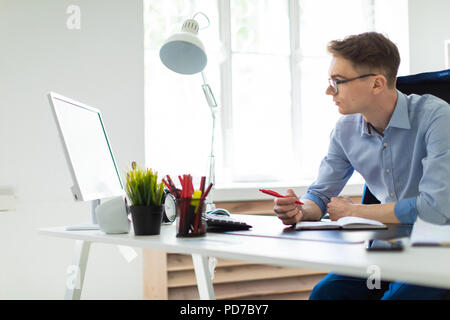 A young man sits in the office at the computer desk, holds a pen in his hand and looks at the monitor. Stock Photo