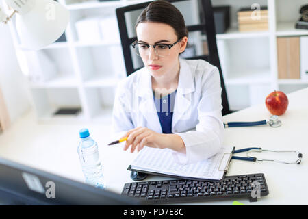 A beautiful young girl in a white robe is sitting at a computer desk with documents and a pen in her hands. Stock Photo