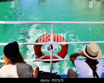 High contrast rear view of 2 young female travellers in sunhats sitting on Lake Komani ferry, Albania, admiring view,a red life belt & no litter sign. Stock Photo