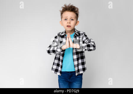 A handsome boy in a plaid shirt, blue shirt and jeans stands on a gray background. The boy folded his arms over his chest. The boy folded his palms against his chest Stock Photo
