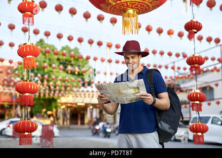 Smiling tourist with a map in the hands stands on the city street and looks at it. He wears a blue polo, crimson hat and a black backpack. Around him  Stock Photo