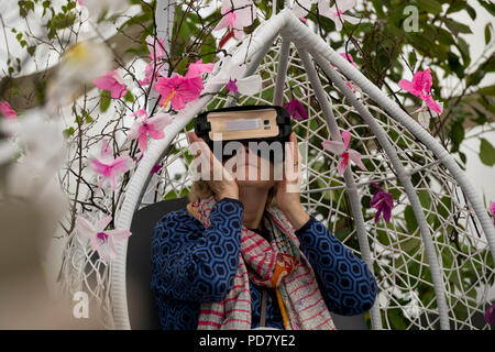 Woman wearing VR headset, sitting in seat among flowers at virtual reality orchid experience - RHS Chatsworth Flower Show, Derbyshire, England, UK. Stock Photo