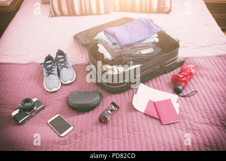 Luggage with pile of clothes, camera, sunglasses and other items on bed Stock Photo