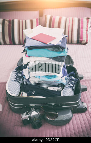 Luggage with pile of clothes, camera, sunglasses and other items on bed Stock Photo