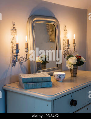 canlde lit wall sconces from The Country Brocante flank a french style mirror above old chest of drawers with fresh flowers Stock Photo