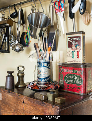 Brass weights and vintage sweet tins sit on an old oak box. Above various kitchen utensils hang from a wall rack Stock Photo