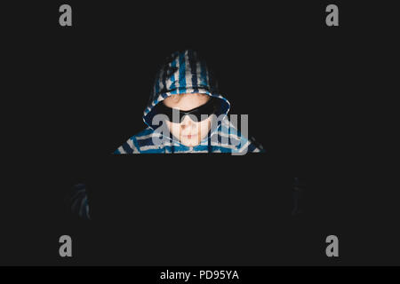 Girl hacker in a hoodie using laptop in a dark room. With sunglasses. Stock Photo