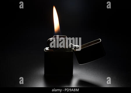 Lighter lit with yellow flame on black background. Vintage stainless steel cigarette lighter with spring flip lid. Stock Photo