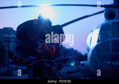 Scary robot going near military helicopter in early morning's downtown. Having big metallic corpus, enormous hand, red diode backlight, looking at cam Stock Photo