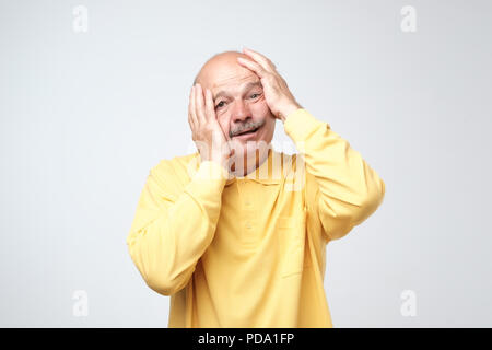 Senior hispanic man in yellow tshirt pulling skin on face with hands. He is scared or tired of stress. I am a little bit crazy concept Stock Photo