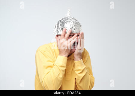 Caucasian mature man in a tin foil hat displeased closing his face with hands. Afraid of radiation or aliens Stock Photo