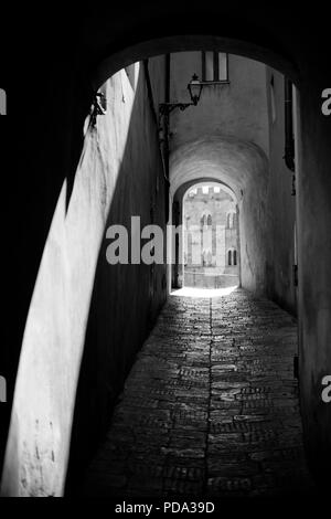 An old passageway with a worn away floor with a view through to the main square in Volterra. Black and white. Stock Photo