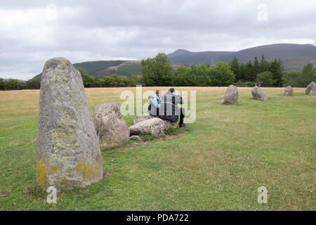 A couple sat on a stone at Castlerigg Stone Circle having a snack with large stone at front and  Lake District mountains in background Stock Photo
