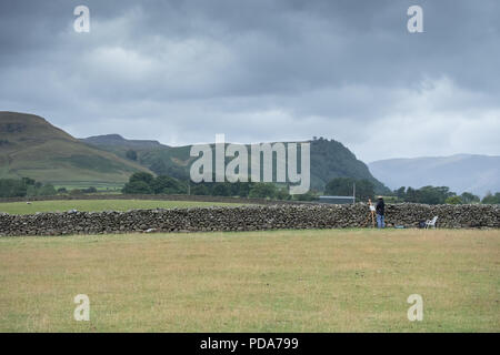 Artist painting his artwork in a field next to Castlerigg stone circle and overlooking the Lake District mountains of Helvellyn in front of stone wall Stock Photo