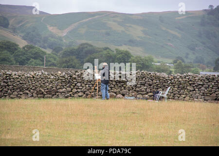 Artist painting his artwork in a field next to Castlerigg stone circle and overlooking the Lake District mountains of Helvellyn in front of stone wall Stock Photo
