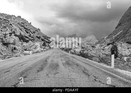 Empty deserted road in black and white leading to menacing stormy, cloudy mountains in the Dolomites, Italy Stock Photo