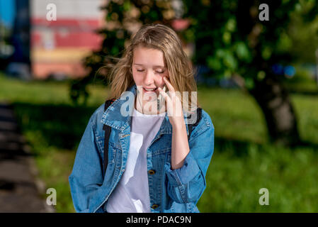 Girl schoolgirl teenager, and in summer in city. Talk on the smartphone, in a denim jacket. Emotionally closes his eyes. Stock Photo